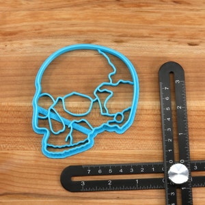 May include: A blue plastic skull-shaped cookie cutter on a wooden surface next to a black ruler.