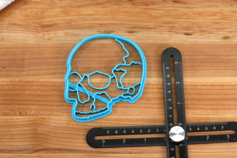 May include: A blue plastic skull-shaped cookie cutter on a wooden surface next to a black ruler.