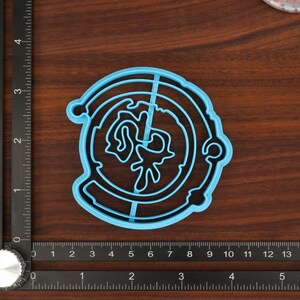 Puede incluir: Un cortador de galletas de pl&aacute;stico azul en forma de c&iacute;rculo con un dise&ntilde;o en el interior. El dise&ntilde;o es una imagen estilizada de un planeta con un anillo alrededor.