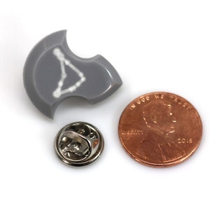 Puede incluir: Un pin de esmalte gris con un contorno blanco de la constelaci&oacute;n de Acuario. El pin es aproximadamente del tama&ntilde;o de una moneda de un centavo de d&oacute;lar estadounidense.