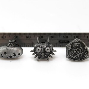 May include: Three silver-coloured metal pins featuring iconic symbols from the Legend of Zelda video game series. The pins are an ocarina, Majora's Mask, and the Hylian Shield.