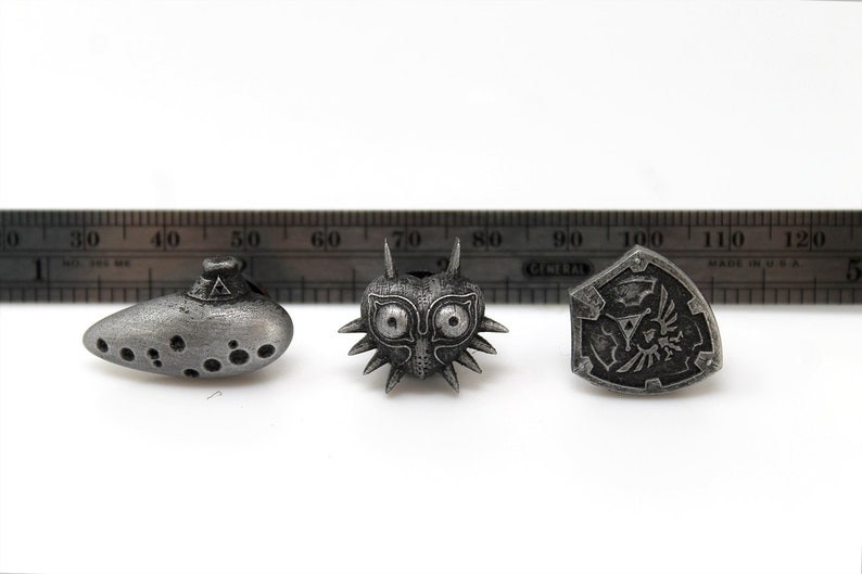 May include: Three silver-coloured metal pins featuring iconic symbols from the Legend of Zelda video game series. The pins are an ocarina, Majora's Mask, and the Hylian Shield.