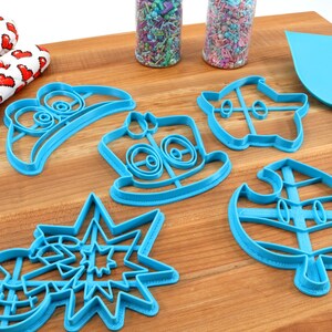 May include: A set of six blue plastic cookie cutters shaped like video game characters. The cutters are on a wooden surface.