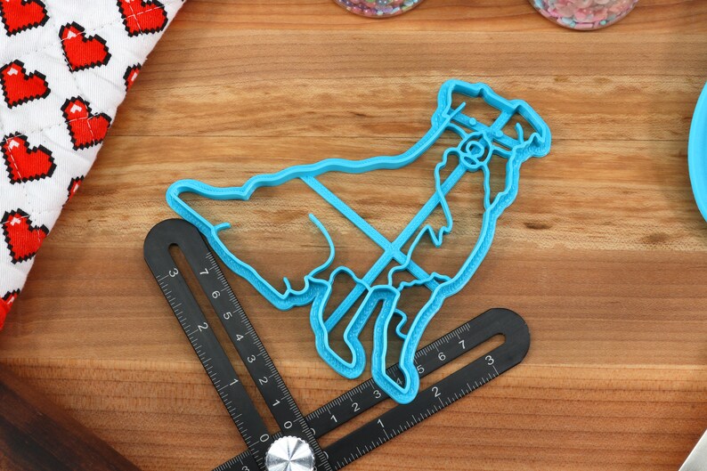 May include: A blue plastic cookie cutter shaped like a goat. The cutter is on a wooden surface with a ruler.