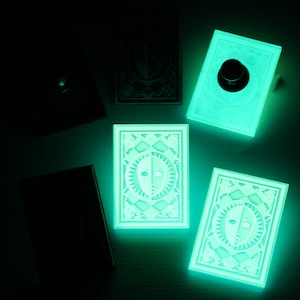 Op de afbeelding: Vijf glow-in-the-dark speelkaarten met een wit ontwerp op een zwarte achtergrond. De kaarten hebben een zon- en maandesign met een gezicht in het midden.