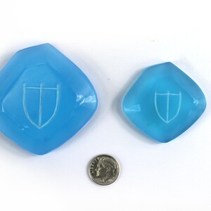 May include: Two light blue glass dishes with a white shield design. The larger dish is about the size of a US dime.