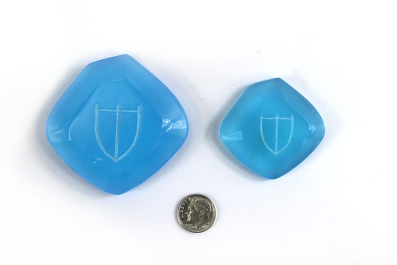 May include: Two light blue glass dishes with a white shield design. The larger dish is about the size of a US dime.