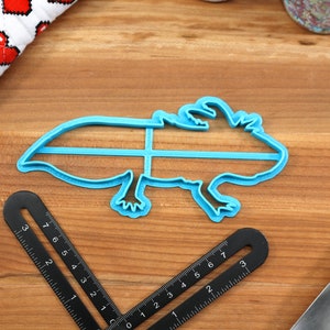 May include: Blue plastic cookie cutter shaped like an axolotl, a type of salamander. The cutter is on a wooden surface with a ruler and a knife.