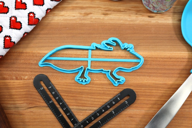 May include: Blue plastic cookie cutter shaped like an axolotl, a type of salamander. The cutter is on a wooden surface with a ruler and a knife.
