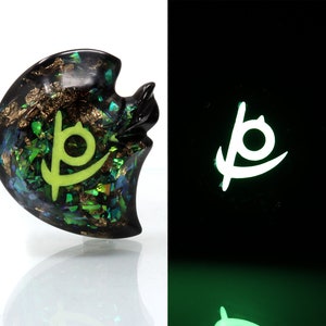 May include: A black resin pendant with a green and gold glitter design. The pendant features a glowing green symbol in the shape of a stylized "P".