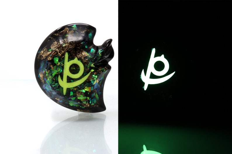 May include: A black resin pendant with a green and gold glitter design. The pendant features a glowing green symbol in the shape of a stylized "P".