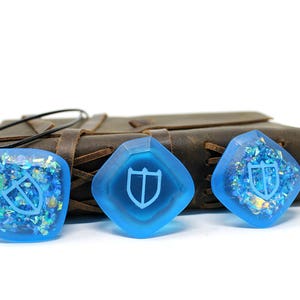 May include: Three blue resin pendants with a white shield design. The pendants are on a brown leather surface.