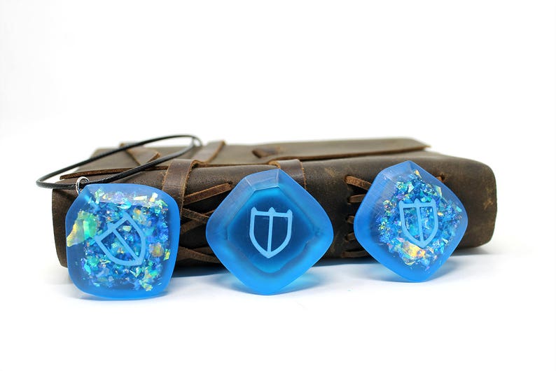 May include: Three blue resin pendants with a white shield design. The pendants are on a brown leather surface.