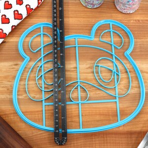 May include: Blue plastic cookie cutter shaped like a cartoon animal with large ears and a grid pattern inside. The cutter is on a wooden surface.