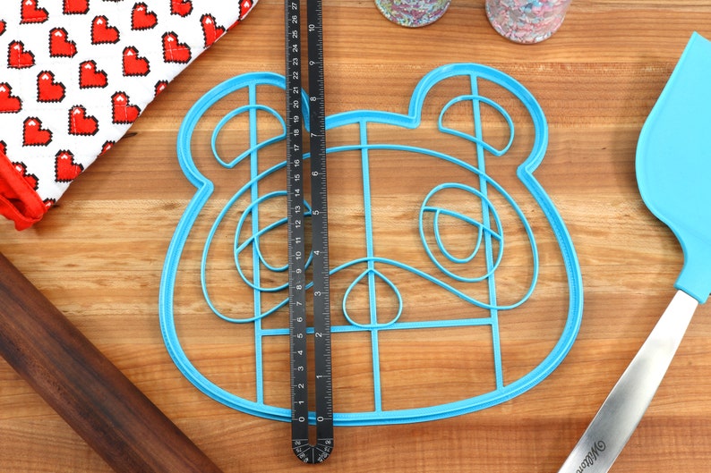 May include: Blue plastic cookie cutter shaped like a cartoon animal with large ears and a grid pattern inside. The cutter is on a wooden surface.