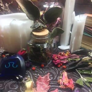 May include: A blue glass object with a symbol on it, surrounded by dried flowers, herbs, and candles. The scene is set on a dark patterned surface.