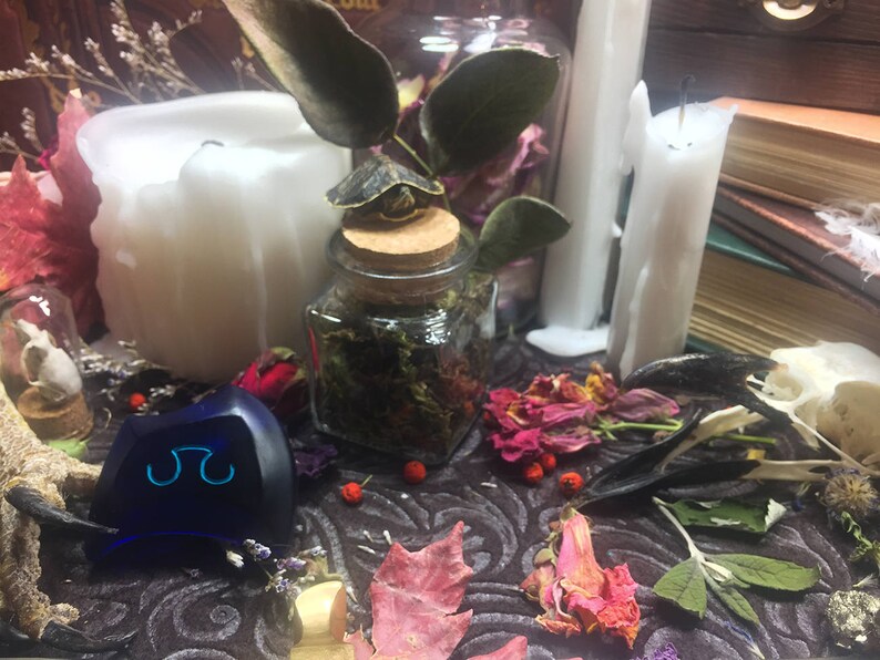 May include: A blue glass object with a symbol on it, surrounded by dried flowers, herbs, and candles. The scene is set on a dark patterned surface.