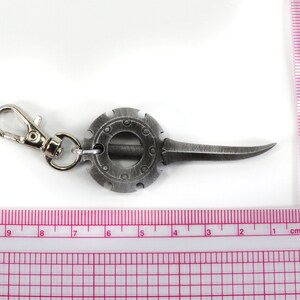 May include: A silver-toned keychain with a circular design and a sharp blade. The keychain has a silver-toned clasp.