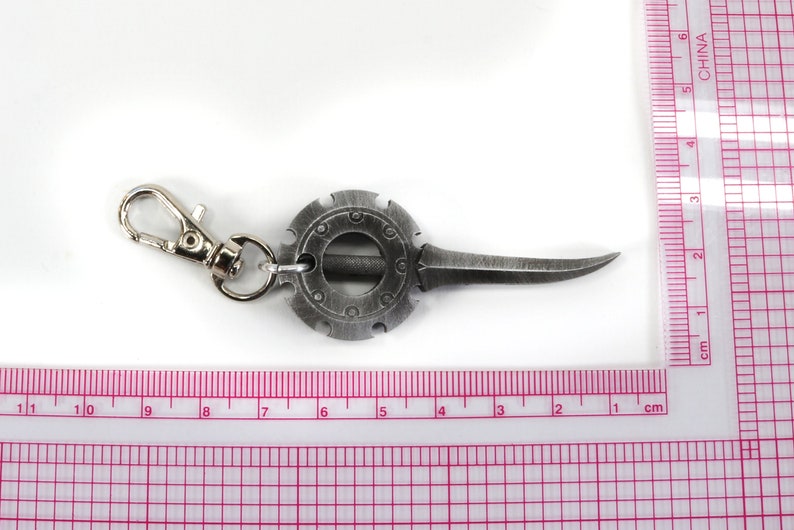 May include: A silver-toned keychain with a circular design and a sharp blade. The keychain has a silver-toned clasp.