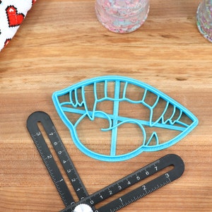 May include: A blue plastic cookie cutter shaped like a mouth with teeth. The cutter is on a wooden surface with a ruler and other baking supplies.