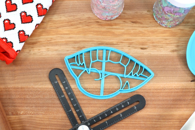 May include: A blue plastic cookie cutter shaped like a mouth with teeth. The cutter is on a wooden surface with a ruler and other baking supplies.