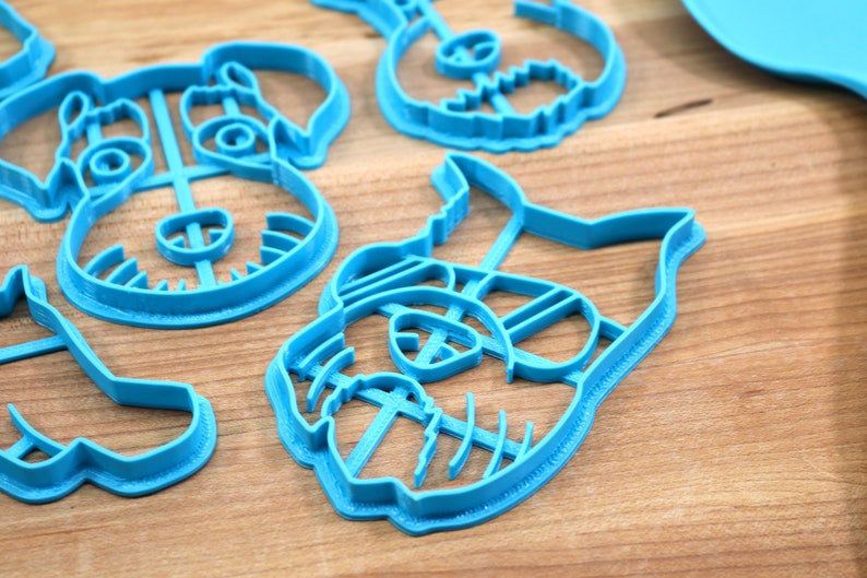 May include: Four blue plastic cookie cutters shaped like dogs. The cutters have detailed features, including ears, eyes, noses, and mouths.
