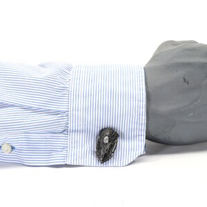 May include: A pair of cufflinks with a black and silver design. The cufflinks are worn on a white dress shirt with blue stripes.