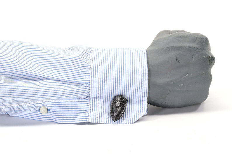 May include: A pair of cufflinks with a black and silver design. The cufflinks are worn on a white dress shirt with blue stripes.