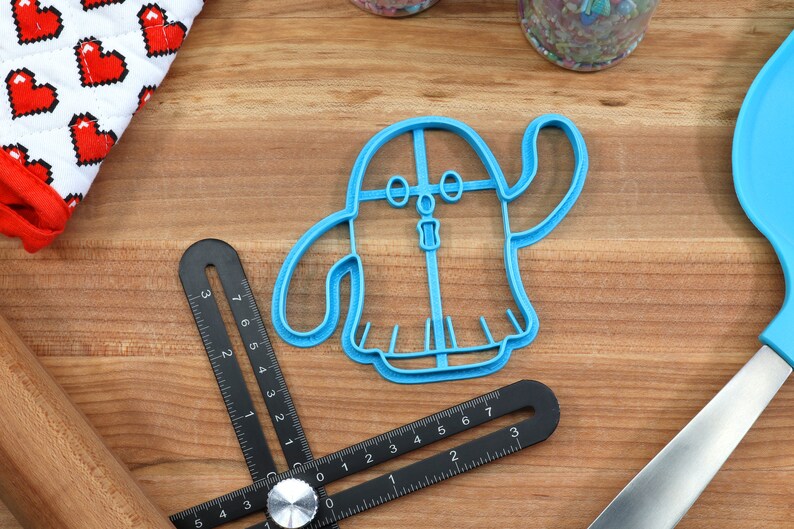May include: A blue plastic ghost-shaped cookie cutter with a friendly face and outstretched arm.