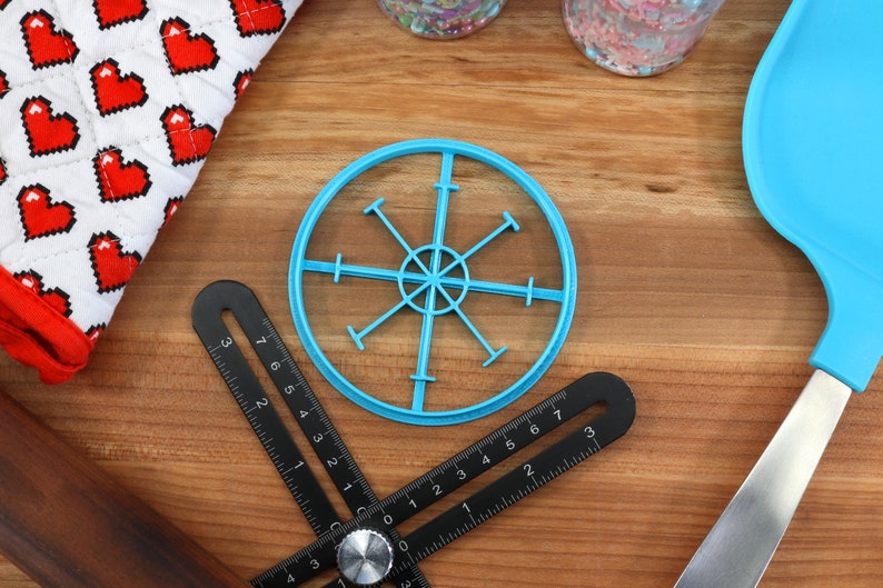 May include: A blue plastic circle cookie cutter with a starburst design. The cutter is on a wooden surface with a ruler and a spatula.