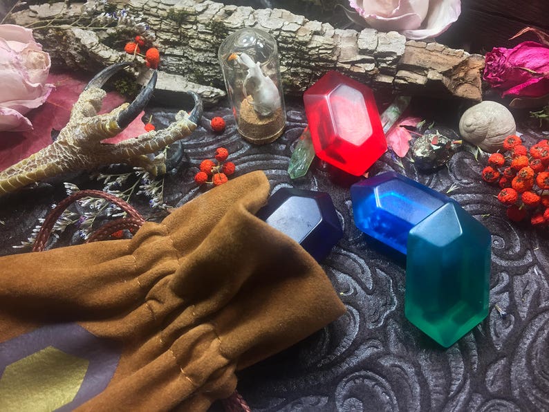 May include: A brown leather pouch with a drawstring closure, filled with four colorful gemstone-shaped objects. The pouch is surrounded by a variety of natural elements, including a bird's foot, berries, and flowers.