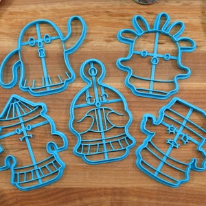 May include: Set of six blue plastic cookie cutters shaped like cartoon characters. The characters have different facial expressions and features, including a ghost, a snowman, a robot, a cactus, and two others.