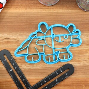 May include: A blue plastic cookie cutter shaped like an axolotl, a type of salamander. The cutter has a simple, cartoon-like design with a smile and large eyes.