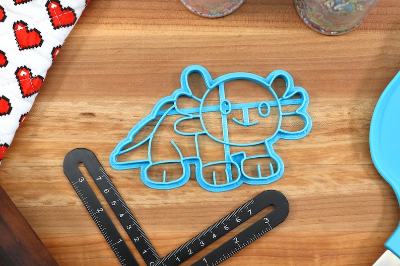 May include: A blue plastic cookie cutter shaped like an axolotl, a type of salamander. The cutter has a simple, cartoon-like design with a smile and large eyes.