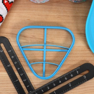 May include: A blue plastic cookie cutter shaped like a heart with a grid pattern inside. The cutter is on a wooden surface with a black ruler.