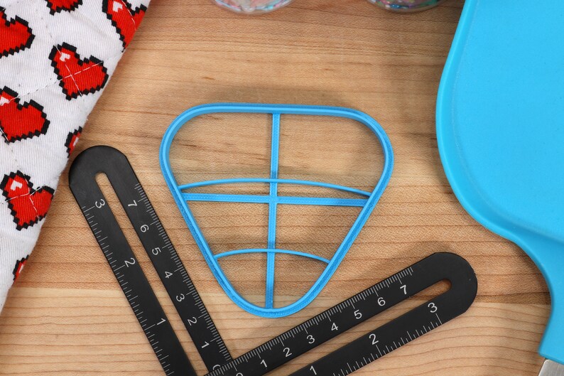 May include: A blue plastic cookie cutter shaped like a heart with a grid pattern inside. The cutter is on a wooden surface with a black ruler.