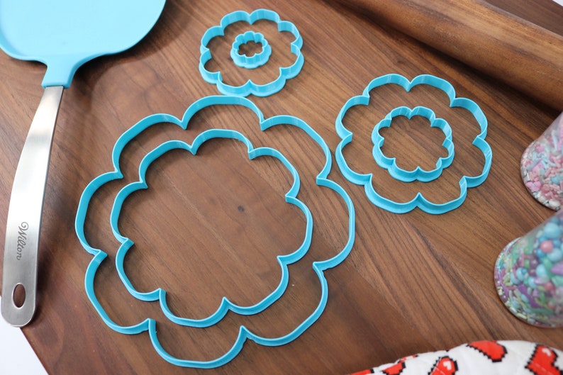 May include: Three blue plastic cookie cutters shaped like flowers, with the largest cutter having two smaller cutters inside it. The cutters are on a wooden surface.