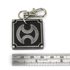 May include: A silver metal keychain with a black square charm featuring a stylized silver symbol. The charm has a silver ring attached to the top for attaching to a keychain.