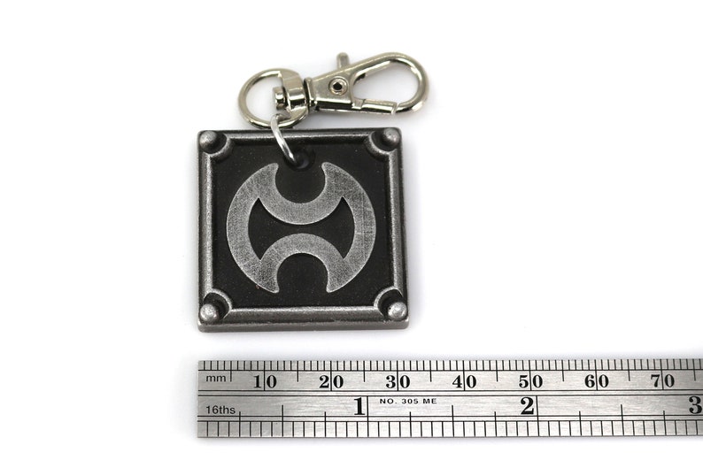 May include: A silver metal keychain with a black square charm featuring a stylized silver symbol. The charm has a silver ring attached to the top for attaching to a keychain.