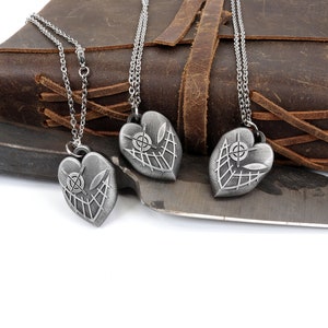 May include: Three silver heart-shaped pendants with a detailed design of a web and a target inside. The pendants are hanging from silver chains on a brown leather background.