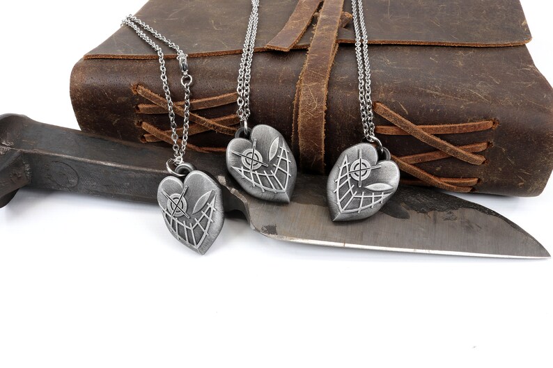 May include: Three silver heart-shaped pendants with a detailed design of a web and a target inside. The pendants are hanging from silver chains on a brown leather background.