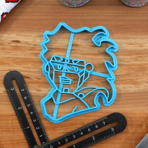 May include: Blue plastic cookie cutter shaped like a cartoon character with spiky hair. The character has a large head and a small body.