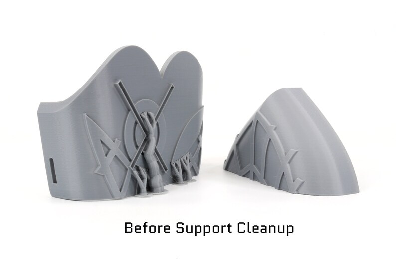 May include: Two gray 3D-printed objects, one with a curved shape and the other with a more complex design featuring a central figure and geometric patterns. The text "Before Support Cleanup" is visible at the bottom of the image.