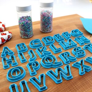 May include: A set of blue plastic alphabet cookie cutters on a wooden surface. The cutters are arranged in alphabetical order. There are two jars of sprinkles in the background.