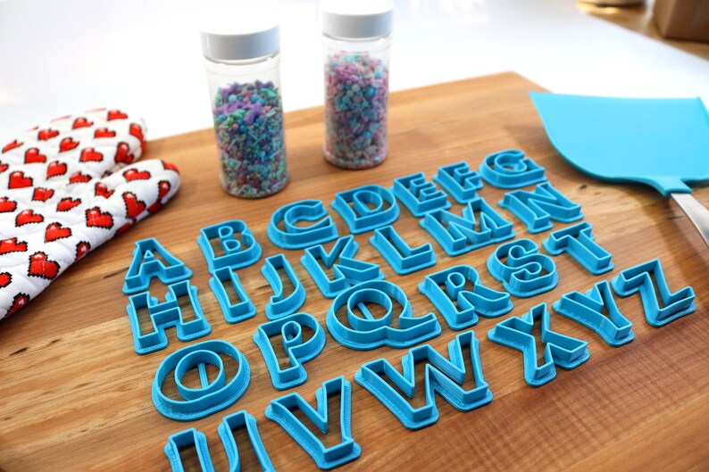 May include: A set of blue plastic alphabet cookie cutters on a wooden surface. The cutters are arranged in alphabetical order. There are two jars of sprinkles in the background.