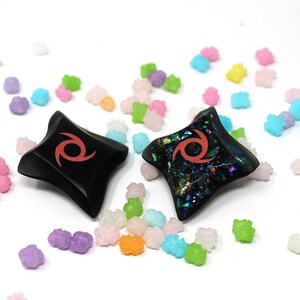 Pu&ograve; includere: Due stelle ninja nere a forma di stella con un design a spirale rosso. Una stella &egrave; nera semplice, l'altra ha una finitura iridescente scintillante. Le stelle sono circondate da spruzzi di caramelle colorate.