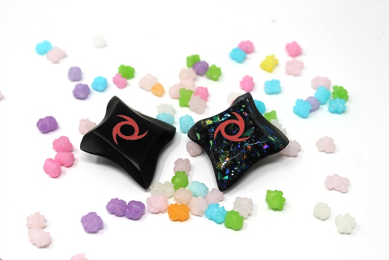 Pu&ograve; includere: Due stelle ninja nere a forma di stella con un design a spirale rosso. Una stella &egrave; nera semplice, l'altra ha una finitura iridescente scintillante. Le stelle sono circondate da spruzzi di caramelle colorate.
