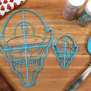May include: Two blue plastic cookie cutters shaped like a cartoon character with a hat and a mustache. The larger cutter is on the left and the smaller cutter is on the right. A blue spatula and a rolling pin are also in the image.