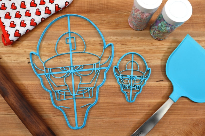 May include: Two blue plastic cookie cutters shaped like a cartoon character with a hat and a mustache. The larger cutter is on the left and the smaller cutter is on the right. A blue spatula and a rolling pin are also in the image.