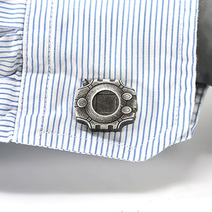 May include: A pair of silver cufflinks featuring a detailed design of a circular device with a square window in the center.